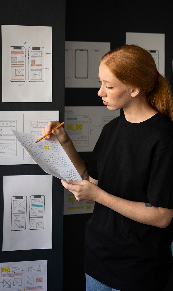Chica revisando el diseño de una web (wireframe) en papel, con una pared llena de bocetos al fondo.